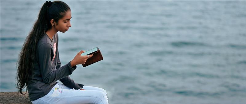 Eine junge Frau sitzt auf einer Steinmauer am Wasser, vertieft in ihr Smartphone, mit Wellen im Hintergrund.