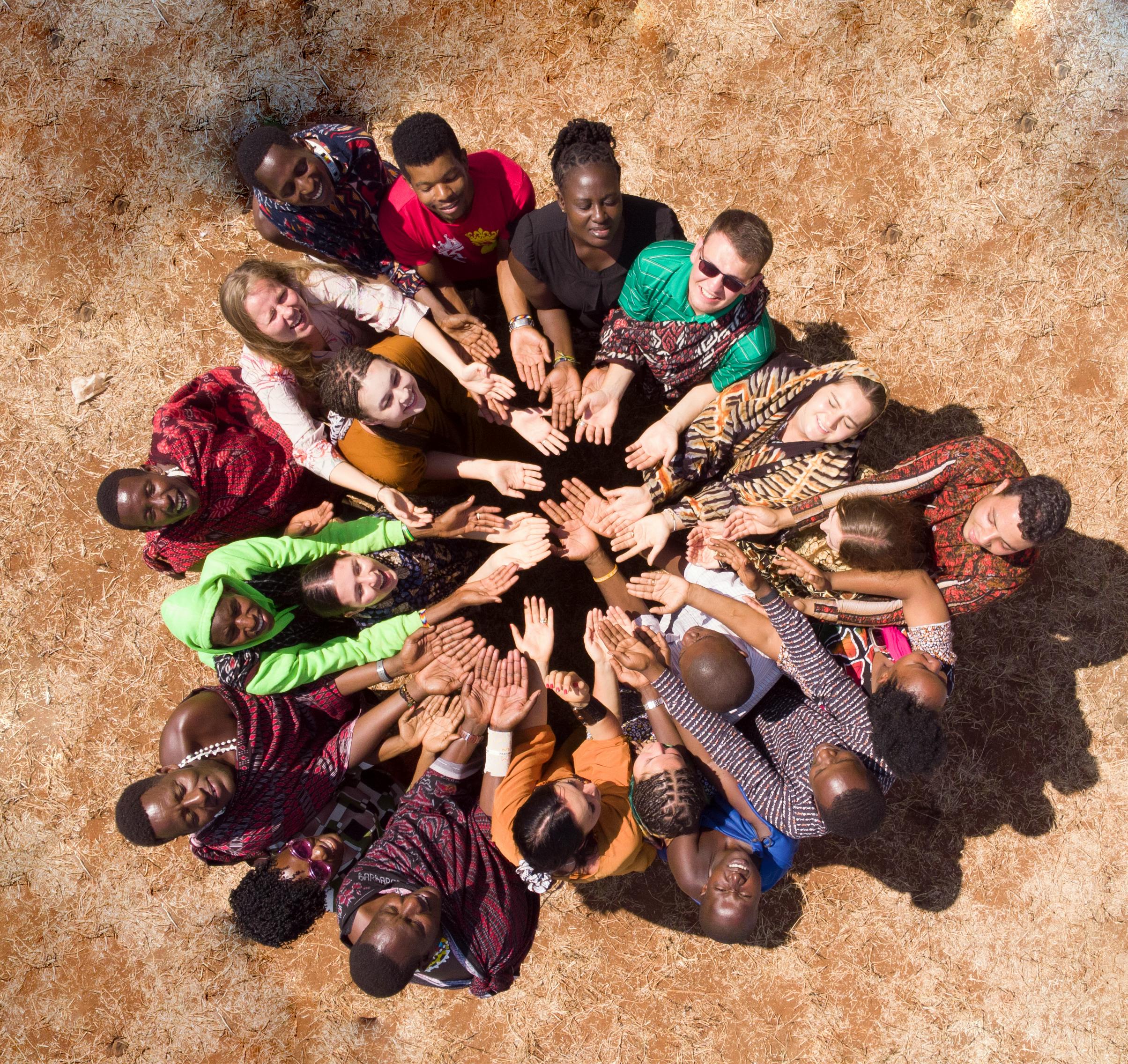 Verschiedenartige Gruppe im Kreis, die Hände zusammengelegt, von oben auf sandigem Boden gesehen.